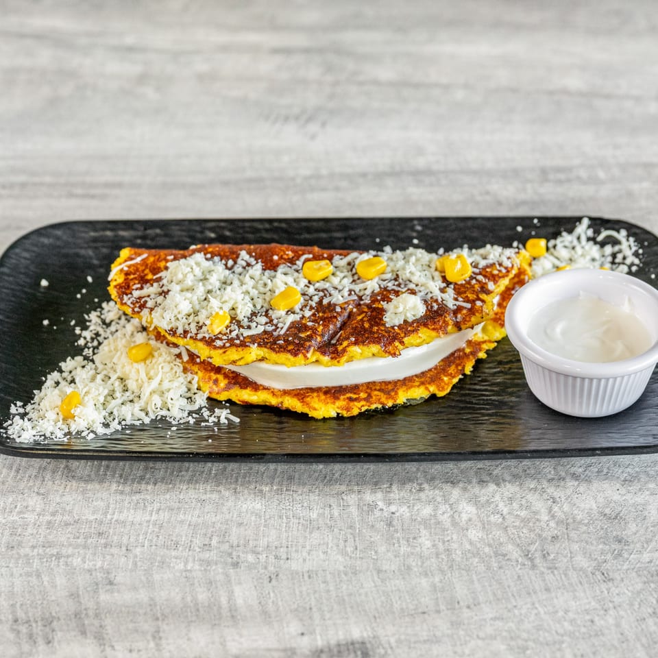 Venezuelan cachapa with cheese and corn, served with a side of nata on a black plate.