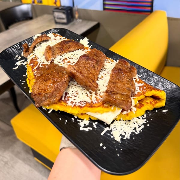 Venezuelan cachapa with steak and shredded white cheese on a black plate, held over a yellow booth.