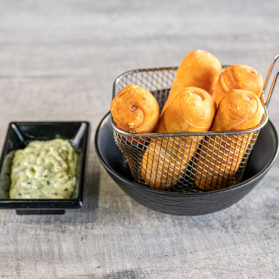 Venezuelan tequeños, golden fried cheese sticks, in a metal basket with a side of green sauce.