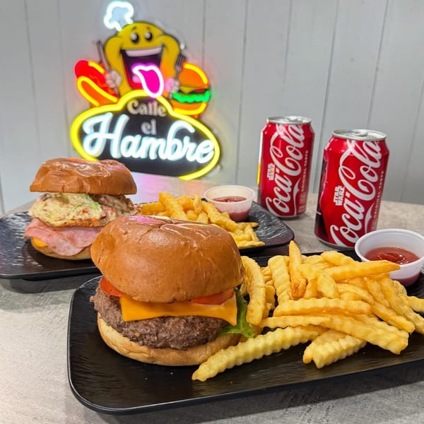 Two Calle El Hambre burgers with fries and two Coca-Cola cans, under a neon sign.