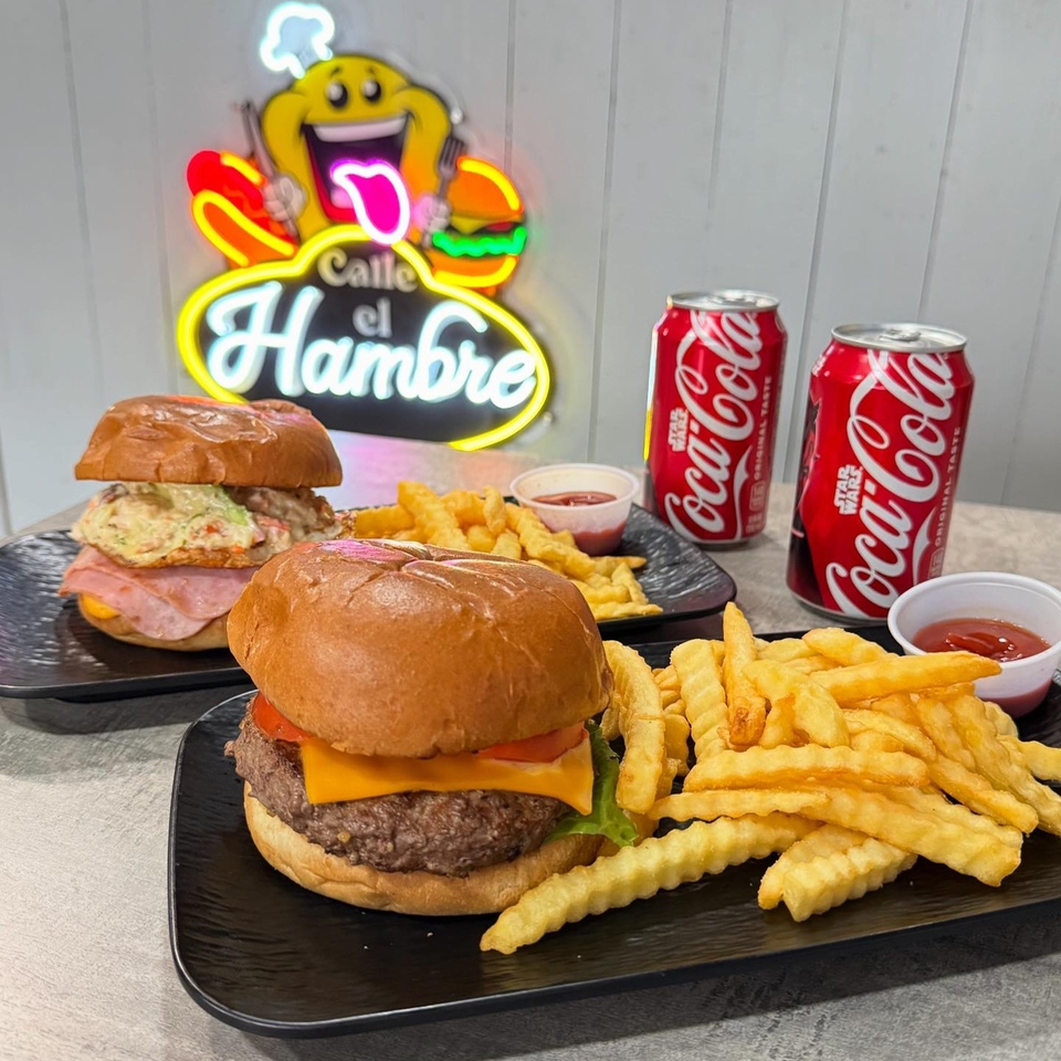 Two Calle El Hambre burgers with fries and two Coca-Cola cans, under a neon sign.