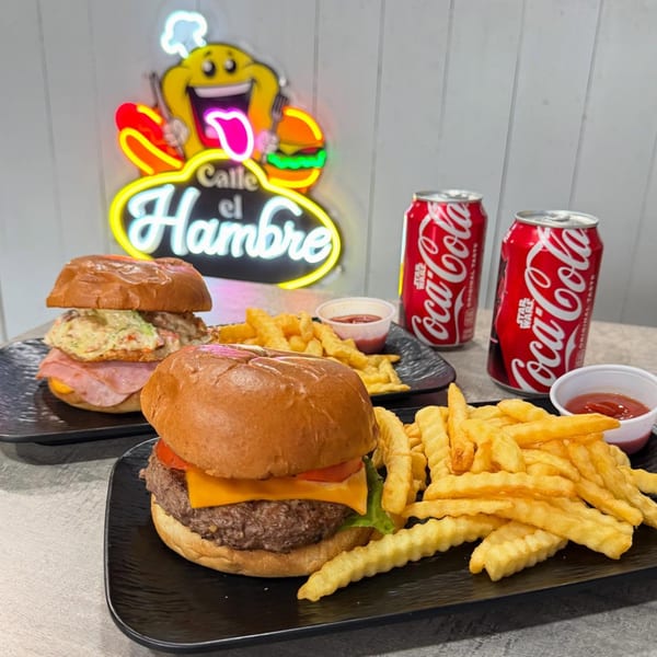 Two Calle El Hambre burgers with fries and ketchup, and two Coca-Cola cans, under a neon sign.